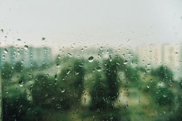 Raindrops on glass close-up against the background of green trees and houses. Cloudy weather and summer storm concept.