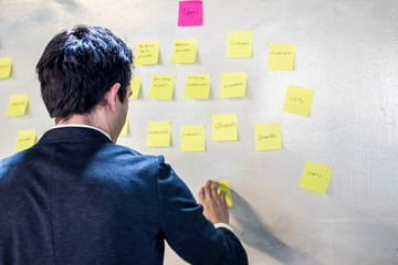 Young Caucasian Male with Dark Hair Writes Notes on Pink and Yellow Sticky Notes on a White Wall in...