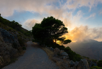 A hiking trail up to Mount Tinoso in eastern Spain. The sun goes down behind a tree. Clouds move between the rocks over the valley. A romantic lighting mood.