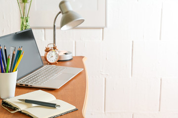 Wooden desk with laptop, notebook, pencils, lamp and tools against a rough white wall, small home office, copy space, selected focus