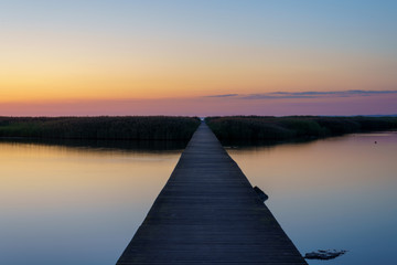 Perspective view of a wooden jetty over sea during summer sunset with reflection in the water.