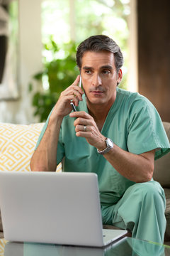 Hispanic Male doctor practicing tele-medicine from his home, using cell phone and laptop computer, Listening to patient on video call 