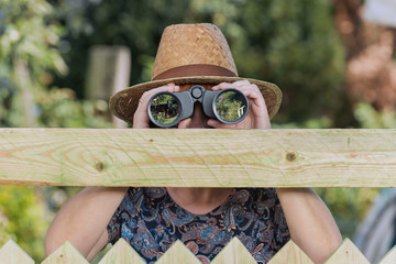 Curious neighbor stands behind a fence and watches with binoculars - obrazy, fototapety, plakaty