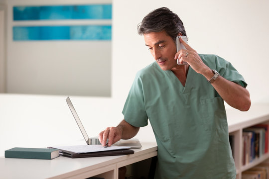 Hispanic Male doctor in his home practicing Telemedicine, talking on cell phone and taking notes 