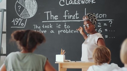 African American female teacher pointing at text on blackboard, speaking to children and answering questions while giving lesson in primary school