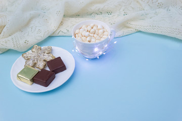 Christmas hot drink and chocolate candies on blue background surrounded by soft knitted scarf. Cozy winter concept. Selective focus, copy space