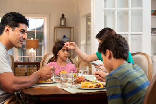 Hispanic Family Around The Dinner Table With The Daughter Feeling Ill, Mom Checking Her Temperature And Checking Cell Phone To Connect With Doctor 