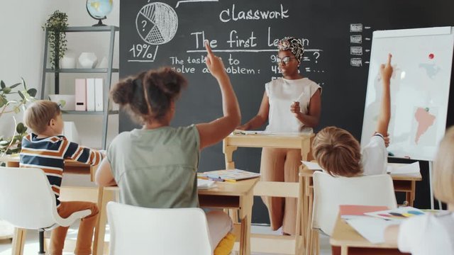 African American female teacher standing beside blackboard and asking a question, then multiethnic kids rising arms up during lesson in primary school