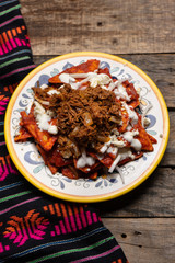 Mexican red chilaquiles with chilorio and cheese on wooden background