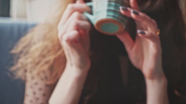 Nice girl takes a cup of hot herbal tea, brings the cup to her lips and drinks from there and laughs while looking at the camera.
