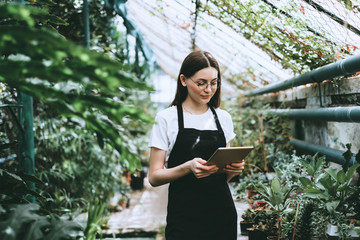 Naklejka na ściany i meble Young woman gardener in glasses and apron with digital tablet working in a garden center for better quality control. Environmentalist using digital tablet in greenhouse.