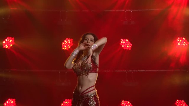 Young female belly dancer performs classical movements of oriental dance. Smooth hand movements. Shot in a dark studio with smoke and red neon lighting. Close up.