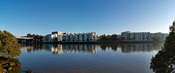 Fototapeta premium Beautiful panoramic view of a river with reflections of modern apartment buildings, deep blue sky and trees on water, Parramatta river, Wilson Park, Silverwater, Sydney, New South Wales, Australia 