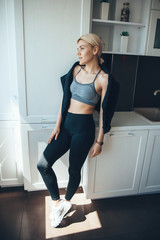 Lovely sporty woman in sportswear is posing at sun in her kitchen at home after doing fitness