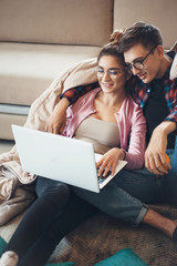 Fototapeta na wymiar Young caucasian couple lying together on the floor near the sofa with a laptop covered with coverlet wearing eyeglasses and smile