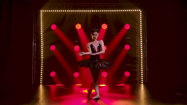 Graceful ballerina in a chic image of a black swan. Young beautiful girl in a black tutu with red sequins and a crown. Perfoming pas with hands. Shot in a dark studio with smoke and red neon lighting.