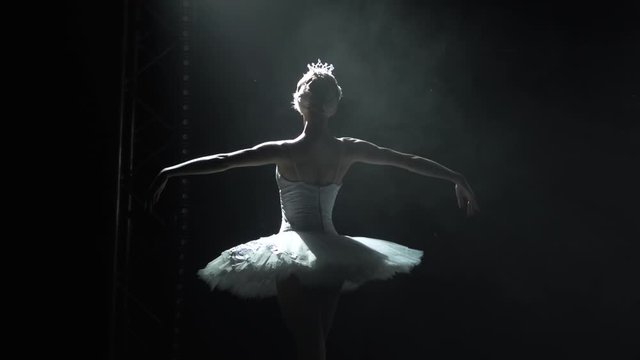 Classical ballet choreography perfoming by young beautiful graceful ballerina in white tutu. Silhouette of a slim figure. Shot in a darkness on spotlights background of studio. Slow motion.