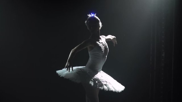 Classical ballet choreography perfoming by young beautiful graceful ballerina in white tutu. Silhouette of a slim figure. Shot in a darkness on spotlights background of studio. Slow motion.