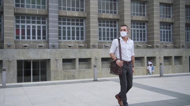 Handsome Caucasian business man office worker carry leather bag walking in downtown district to office building. Businessman wearing face mask during protect covid-19 virus pandemic and air pollution.