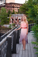 Beautiful blonde woman in pink strapless dress stands on walk way lined with greenery and abandoned building behind her