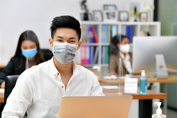Portrait of Asian man office worker wearing face mask working in new normal office and doing social distancing during corona virus covid-19 pandemic - Powered by Adobe