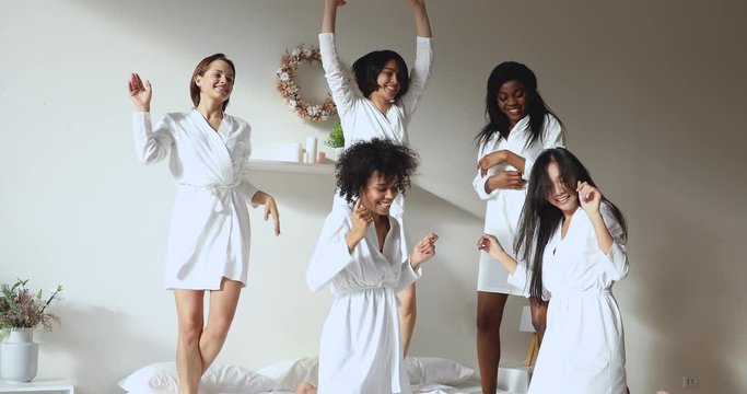 Overjoyed young multiracial diverse beautiful women in white bathrobes dancing, having fun jumping on bed, enjoying spa day together or celebrating bachelorette hen birthday girlish party in hotel.