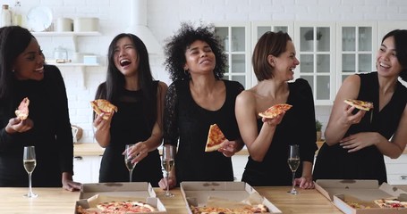 Happy young festive mixed race women in black dresses eating pizza, drinking champagne, celebrating bridal shower together. Excited diverse girls best friends enjoying thematic birthday party at home.