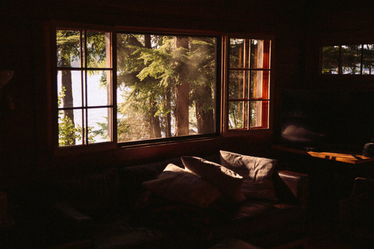 Window With Light Coming Through And Illuminating A Living Room With A Couch And Pillows; Trees, Lake, Water, And Sun Outside The Window