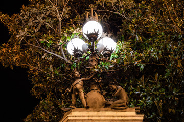 Two bronze angels with a sword and playing the drum at the foot of a lamppost with three lights lit under the leaves of a tree next to the Buenavista Palace Gardens