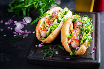 Hot dog with sausage, cucumber, tomato and red onion on dark background.