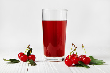 Transparent glass of fresh cherry juice and some fresh cherries near on white wooden boards against light wall.