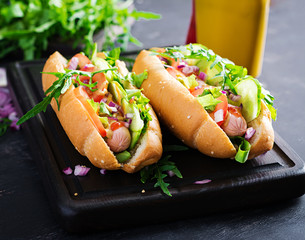 Hot dog with sausage, cucumber, tomato and red onion on dark background.