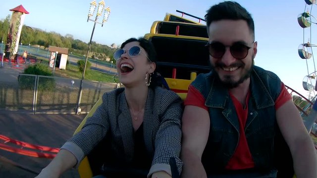 Selfie video of couple riding the roller coaster. Hipster man and stylish woman have fun at amusement park rides. Friends extreme riding, laughing and screaming of excitement, adrenalin.