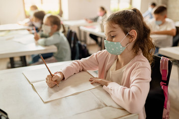 Schoolgirl with protective face mask writing in a notebook during a lecture in the classroom.