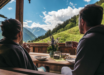 Couple sitting outside of a mountain hut, eating breakfast and enjoying the scenery - obrazy, fototapety, plakaty