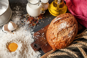 On the board is a loaf of rye bread. Nearby is a bottle of milk and butter. Items are located on a black background. Flour is scattered on the table.