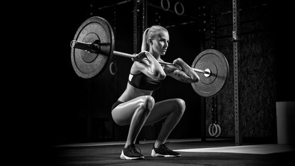 Naklejka na ściany i meble Indoors shot of muscular young woman doing squats with barbell in a gym. Female bodybuilder workout Confident young blonde woman doing crossfit workout in gym Black and white photo.