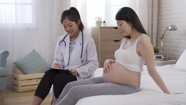 asian pregnant woman sitting on the edge of bed is answering the obstetrician’s questions. chinese female doctor is having a pleasant conversation with her patient during home visit.