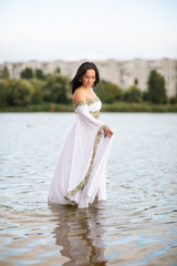 Fototapeta na wymiar Young european woman with brown hair dressed a long white wedding dress stays in the river