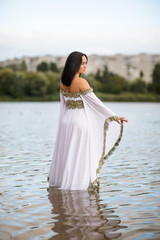 Young european woman with brown hair dressed a long white wedding dress stays in the river