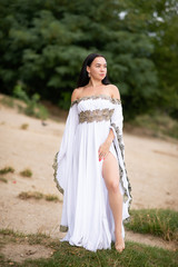 Young european woman with brown hair dressed a long white wedding dress stays near the river