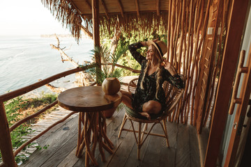 Attractive Woman in Hat  sitting on the chair and drinking coconut cocktail at beautiful location with sea view. Fresh tropical fruits and coconut for breakfast on table.