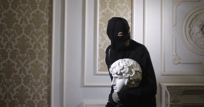 Robbery of the museum. A man in black clothes, a mask and gloves stealing  the sculpture. A funny and stupid robber sneaking up with a statue in hand. Security system concept. 