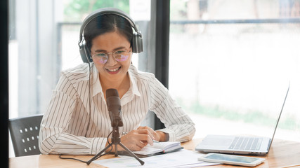Asian women podcaster podcasting and recording online talk show at studio using headphones, professional microphone and computer laptop on table looking at camera for radio podcast