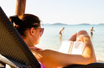 Woman on sun bed on the beach reading a book. People in background walking into sea. Croatian...