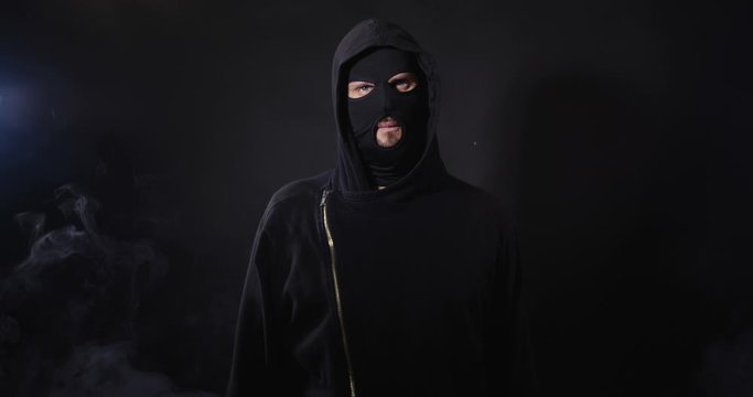 Portrait of robber in black clothes and mask looking aggressive to camera on black studio background with smoke. Security system concept.  4K 50 fps slow motion
