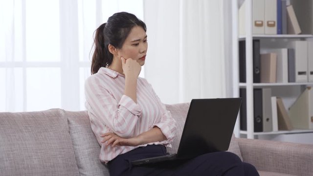 female chief editor copyediting manuscript on laptop and thinking about how to better it. asian woman stroking her chin considering word choice. work from home concept