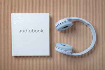 White headphones and a white book on the blue and beige background. Audio book reading online.