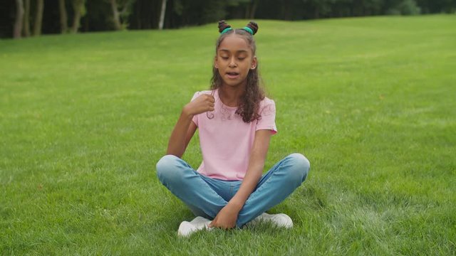 Positive confident attractive preadolescent african american blogger girl sitting cross-legged on green park lawn, broadcasting video content for vlog, sharing with her audience online in nature.