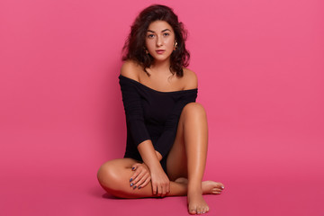 Portrait of cute female wearing black bodysuit, sitting on floor against rose wall, looks at camera, has dark wavy hair, posing with stylish earrings.